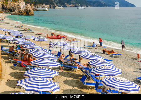 Der Sandstrand von Monterosso, Cinque Terre Italien, mit Sonnenschirmen, Sonnenanbeter, das Ligurische Meer und Boote entlang der felsigen Küste. Stockfoto