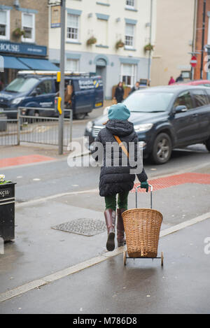 Wetter: Bridport, England, UK Freitag, den 9. März 2018 eine Frau mit einem blauen Hut und einem Korb Warenkorb geht auf ein Auto in den Regen auf Bridport High Street. Die trüben und nassen Bedingungen über das Wochenende mit einer Verbesserung am Montag voraussichtlich fortsetzen. Credit: PQ Images/Alamy leben Nachrichten Stockfoto