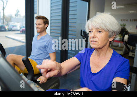 Ältere Frau Spinnen auf Fitness Bikes mit Personal Trainer Stockfoto