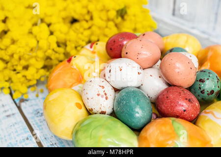 Bunte Ostereier auf einem weißen Teller in der Form der Herzen und Blumen von Mimosa auf einem blauen Holztisch. Close Up. Stockfoto