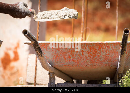 Nur mit der Hand der Bauarbeiter auf der Baustelle, nassen Zement auf einer kelle von seiner Schubkarre, Gebäude Betonwand Struktur mit Stahlstangen. Stockfoto