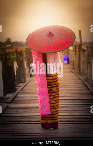 Frau Myanmar Holding traditionellen roten Regenschirm auf U Bein Holzbrücke bei Sonnenaufgang in Mandalay, Myanmar. Stockfoto