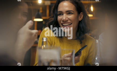 Schöne Hispanic Frau nutzt Smartphone bei der Unterhaltung und Spaß mit Ihren Freunden in der Bar. Sie Lachen, Scherzen, Getränk in der stilvollen Bar Hipster Stockfoto