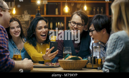 Schöne Hispanic Frau zeigt interessante Sachen auf Ihrem Smartphone zu Ihren Freunden während Sie gute Zeit in Bar. Sie Lachen, Scherzen, Getränk in der Bar Stockfoto