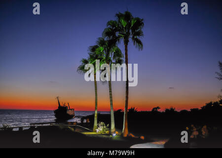 Schiffbruch bei Sonnenuntergang Zeit in Paphos, Zypern mit drei großen Palmen, von der Basisstation leuchtet. Im Vordergrund Silhouette. Stockfoto