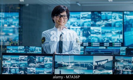 In den Sicherheitsraum Officer Monitore Bildschirme für verdächtige Aktivitäten, er lächelt in die Kamera. Bildschirme zeigen internationale Logistik. Stockfoto