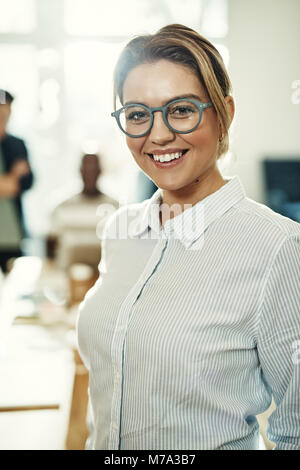 Junge Geschäftsfrau mit Brille und lächelnd, während er sich in einem Büro mit den Kollegen hinter ihr zu arbeiten Stockfoto