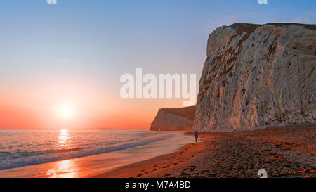 Sunset at Durdle Door Strand. Lone fischer Spaziergänge zu den Weißen Klippen, Rot der untergehenden Sonne. Die Leute stehen auf der Spitze einer Klippe. Stockfoto