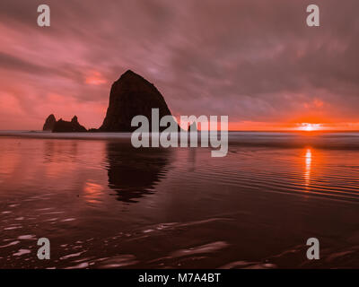 Sonnenuntergang an der Küste von Oregon. Haystack Rock Silhouette spiegelt sich im Wasser bei Cannon Beach. Flammend roten Nachleuchten als Sonne am Horizont. Stockfoto