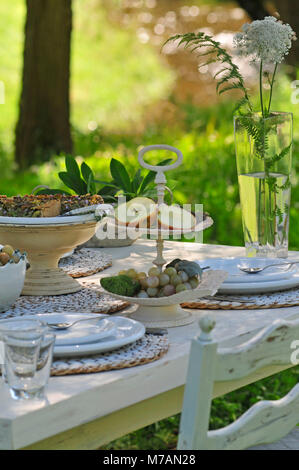 Mangold Quiche und Etagere mit in Scheiben geschnittenen Apfel auf weiß gedeckten Tisch, in der Hintergrund verschwommen Wiese und Bach Stockfoto