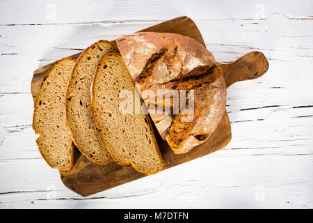 Laib Brot, teilweise geschnitten, auf hölzernen Breadboard und weißen Holzmöbeln Hintergrund, Ansicht von oben, close-up. Stockfoto