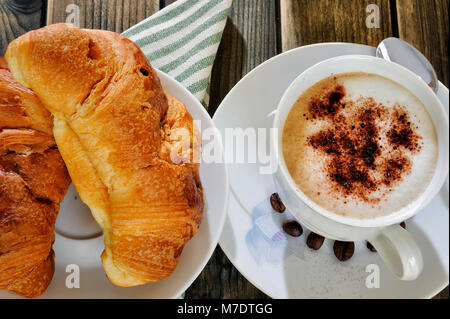 Nahaufnahme einer Tasse Kaffee mit Milch und Croissants auf einem antiken Holztisch. Stockfoto