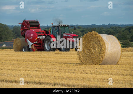 Pressen von Stroh in Bauernhof Feld, Landwirt arbeitet und Laufwerke, Red Tractor Pulling Rundballenpresse (große Ballen gerade freigegeben) - Whixley, North Yorkshire, England, UK. Stockfoto