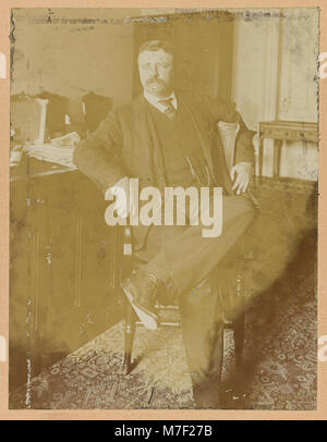 Ein formelles Porträt von Theodore Roosevelt, das ihn mit überkreuzten Beinen an einem Schreibtisch zeigt. Dieses dreiviertel lange Bild fängt den Präsidenten in einem Moment der stillen Reflexion oder Arbeit ein, symbolisch für seine Führung und Persönlichkeit. Stockfoto