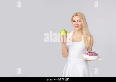 Schöne blonde Frau in weißem Kleid zwischen leckeren Kuchen und frischen Apfel wählt. Lecker oder gesundes Essen Stockfoto