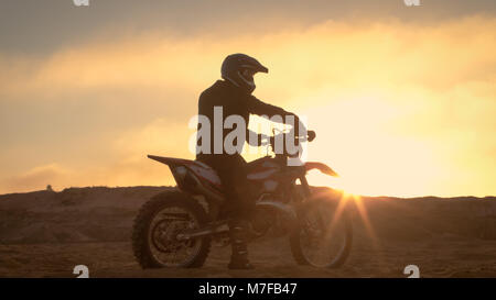 Professionelle FMX Motorradfahrer ruht auf seinem Fahrrad und blickt auf harte Sandy Off-Road Gelände. Sun Einstellung ist. Stockfoto