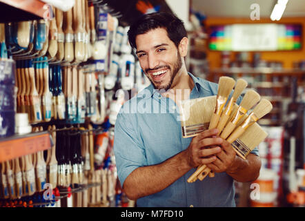 Junger Mann, der unter den Regalen in Farbe Speicher Auswahl von Pinseln Zufrieden Stockfoto