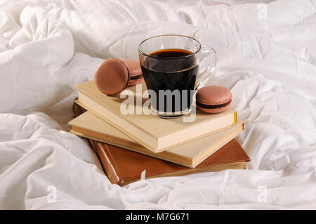 Morgendlichen Tasse Kaffee mit Schokolade Kuchen Makronen, auf einem Stapel Bücher im Bett. Stockfoto