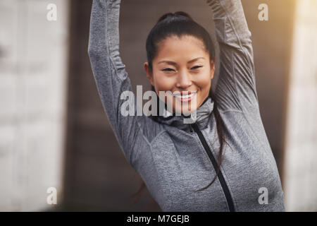 Passen junge asiatische Frau in Sportkleidung lächelnd, während außerhalb recken ihre Arme vor der Ausübung Stockfoto