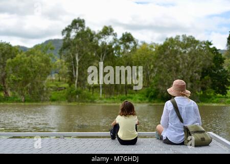 Mutter und Tochter sitzen am Pier mit Blick auf Wasser und Wald sich über Themen, Apex Park, Riverway Drive, Condon, QLD, Australien Stockfoto