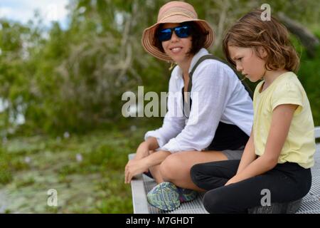 Mutter und Tochter sitzen am Pier mit Blick auf Wasser und Wald sich über Themen, Apex Park, Riverway Drive, Condon, QLD, Australien Stockfoto