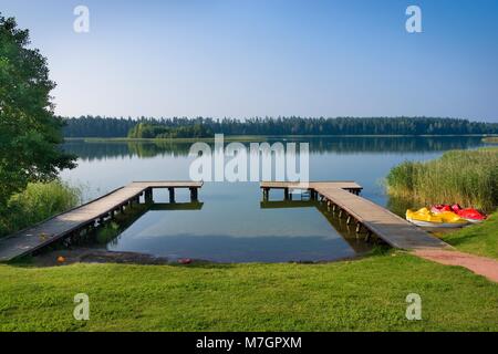 Tretboote durch leere Holzsteg vertäut am Ufer des Sees mit der Insel im Hintergrund, Blizno See, Polen Stockfoto