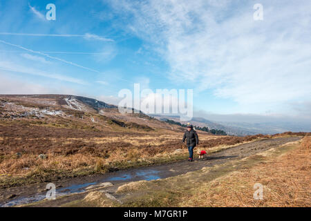 Frau in einem Bobble hat man ein kleines Spielzeug Rasse Hund in einem roten Mantel entlang Ilkley Moor an einem kalten, aber sonnigen Tag mit Remants Schnee Stockfoto