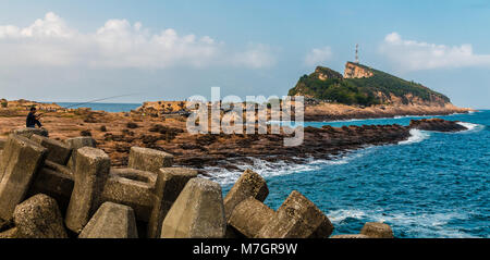 Ein Mann angeln auf einen Wellenbrecher Struktur. Im Hintergrund ist der Gueitou Berg Der yehilu Geopark Kap. Im August 2014, Taiwan. Stockfoto