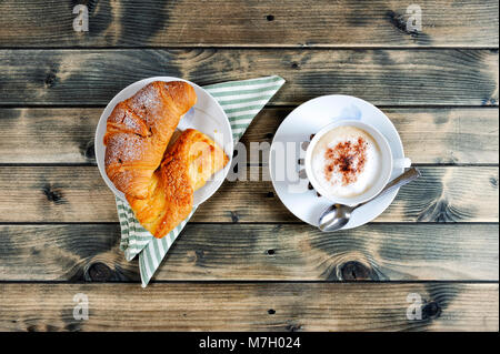 Blick von oben auf eine Tasse Kaffee mit Milch, Croissants und Brioche auf einem antiken Holztisch. Stockfoto