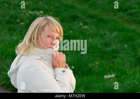 Seite im freien Kopf und Schultern eines Blonde reife Frau auf der grünen Wiese die Hände am Revers und glücklich lächelnd in die Kamera. Stockfoto