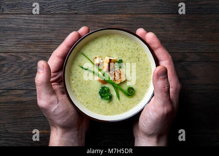 Vegaterian winter Split Pea Kurkuma Suppe mit Croutons und Grün in lila Keramik Schüssel durch die Hände des Menschen holden auf dunklem Hintergrund, Stockfoto