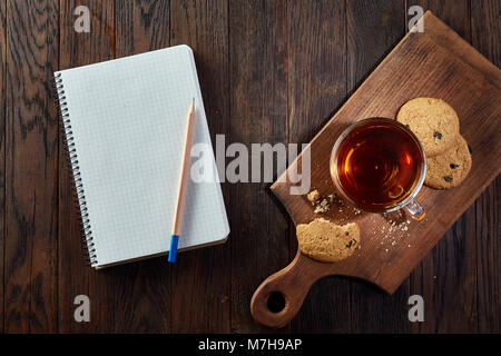 Tasse Tee mit Chocolate Chips cookies, Arbeitsmappe mit in es und einen gelben Bleistift auf einem hölzernen Schneidebrett über vintage Hintergrund tun, oben vie Stockfoto