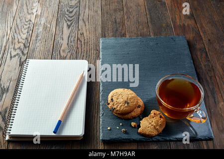 Tasse Tee mit Chocolate Chips cookies, Arbeitsmappe mit in es und einen gelben Bleistift auf einem hölzernen Schneidebrett über vintage Hintergrund tun, oben vie Stockfoto