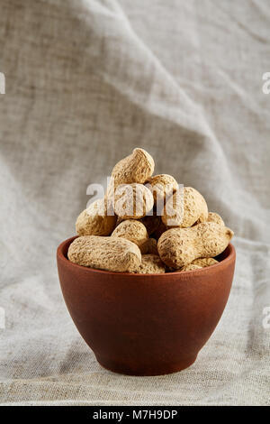 Ungeschälte Erdnüsse in Ton Keramik Schüssel closeup, selektiver Fokus, senkrecht. Gesunde und nahrhafte Zwischenmahlzeit. Köstliche Zutat für Ihr jeden Tag kochen Stockfoto