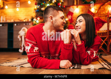 Glücklich Liebe Paar auf dem Boden liegt, Weihnachtsferien. Weihnachten Feier, freundliche Mann und Frau in Rot Pullover, Tanne mit Dekoration auf Backgr Stockfoto