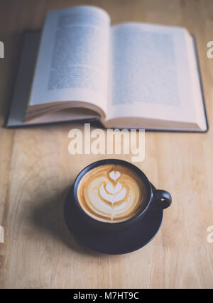 Latte Art Kaffee in schwarzer Schale mit offenem Buch auf Holz Tisch im Café Stockfoto