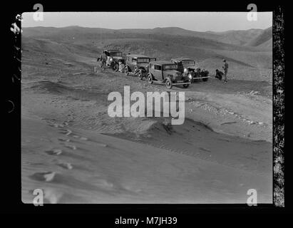 Dieses Bild zeigt Autos, die durch eine Wüstenlandschaft navigieren und den starken Kontrast zwischen modernen Verkehrsmitteln und der riesigen, kargen Umgebung hervorheben. Stockfoto