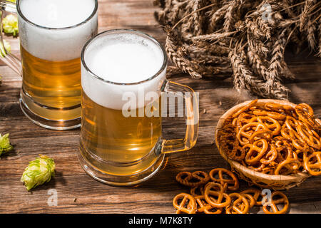 Becher schaumiges Bier, Hopfen, Brezeln und Weizen auf hölzernen Tisch Stockfoto