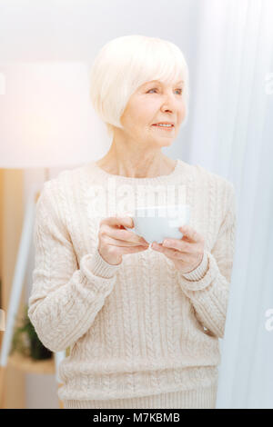 Nachdenklich ältere Frau stand in der Nähe des Fensters und halten einer Tasse Stockfoto