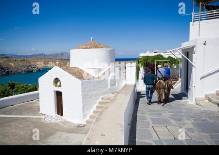 Menschen, die auf Eseln im traditionellen griechischen Dorf, Agios Georgios Hostos Kirche auf der linken Seite, Lindos, Insel Rhodos, Griechenland Stockfoto