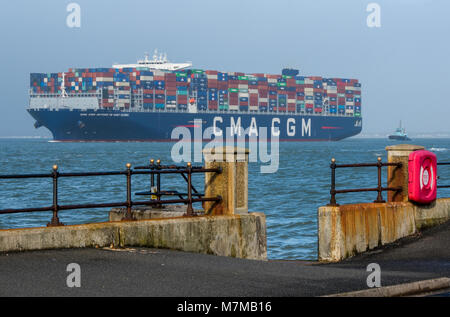 Eine große massive Container schiff betreten oder verlassen den Hafen von Southampton Docks. Schlepper hinten an ein riesiges Schiff schleppen und Abschleppen Behälter angebracht ist. Stockfoto