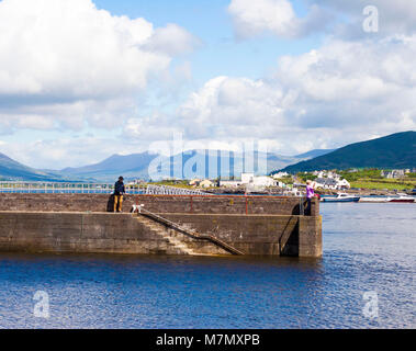 Mann, der seinen Hund am Ende der Pier, Hafenmauer, orange Kanu, vor einer atemberaubenden Landschaft Hintergrund, Ring of Kerry Kerry Irland Stockfoto