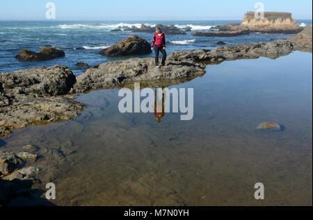 Eine Frau steht auf Felsen im Meer. Sie schaut auf das Ufer, während ein Lächeln auf den Lippen. Stockfoto