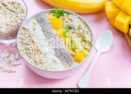 Mango, Banane, Ananas und Haferflocken Smoothie in der Schüssel mit Zutaten in der Nähe auf der Pastell rosa Hintergrund Stockfoto