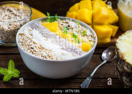 Mango, Banane, Ananas und Haferflocken Smoothie in der Schüssel mit Zutaten, die auf dem dunklen Hintergrund Stockfoto