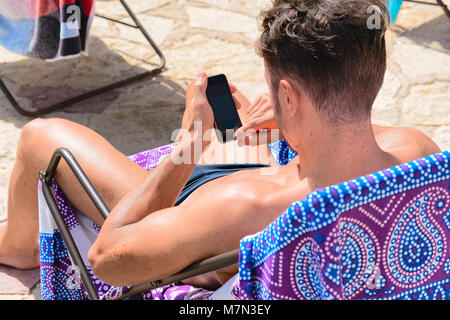 Junger Mann auf sein Smartphone am Strand suchen, Scopello, Castellammare del Golfo, Sizilien, Italien Stockfoto