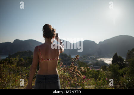 Braungebrannte Mädchen steht auf einem Hügel und macht ein Foto von der schönen Landschaft. Rückansicht des unkenntlich junge Frau. Tropisches Reiseziel Stockfoto