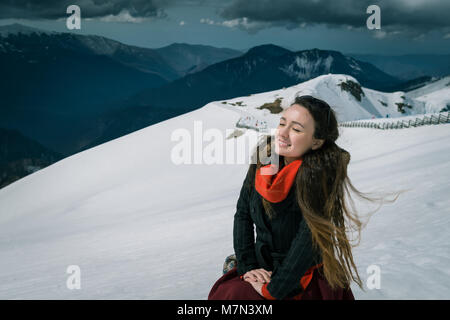 Junge Frau sitzt auf der Spitze der schneebedeckten Berge im Skigebiet. Trendy Traveler bietet einen wundervollen Tag und nimmt ein Sonnenbad. Stockfoto