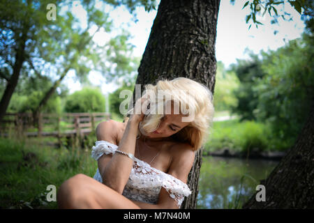 Junge Frau lächelt und setzt sich auf Gras unter den großen Baum in der Nähe des Flusses im Park. Schlanke blonde Mädchen mit schönem Haar in weißem Kleid auf der Natur Stockfoto