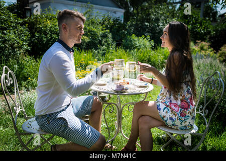 Mann mit Freundin sitzt an stilvollen Tisch mit Süßigkeiten in den grünen Garten. Glückliches Paar Getränke Kaffee im Hof. Junge Frau mit Freund mit Cups Stockfoto